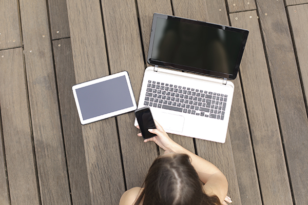Woman using multiple devices phone laptop and tablet lying in a wood bench in a park
